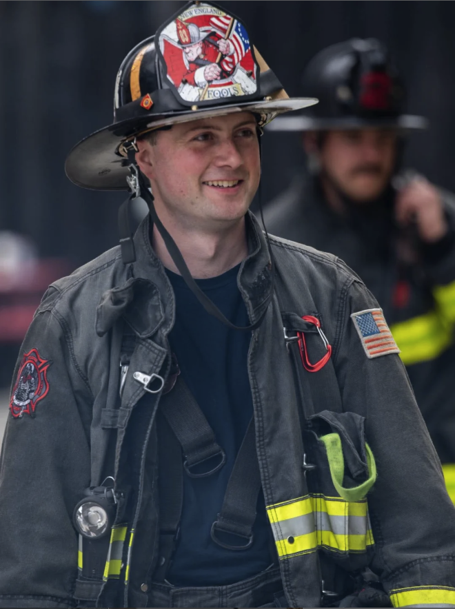 Marty Parker Jr. smiling in full turnout gear and helmet with a New England Fools front shield, American flag shoulder patch, and reflective stripes.